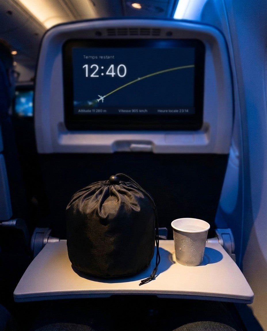 Cabine d'avion tamisée de nuit, l'écran du siège affiche 12:40 de temps restant, le coussin Calé rangé dans sa pochette fermée sur la tablette